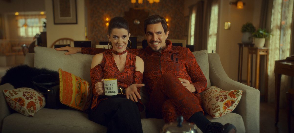 Mary Holland and Josh Segarra, wearing flashy red outfits, smile on a couch in The Big Door Prize. Holland is holding a mug that says “Whiskey while you work.”