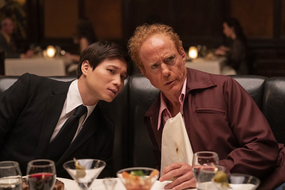 Robert Downey Jr. and Hoa Xuande speak at a dinner table in The Sympathizer. Hoa Xuande wears a nice black suit with a black tie, while Downey Jr., disheveled and with red hair, wears a red collared jacket with a napkin as a bib.