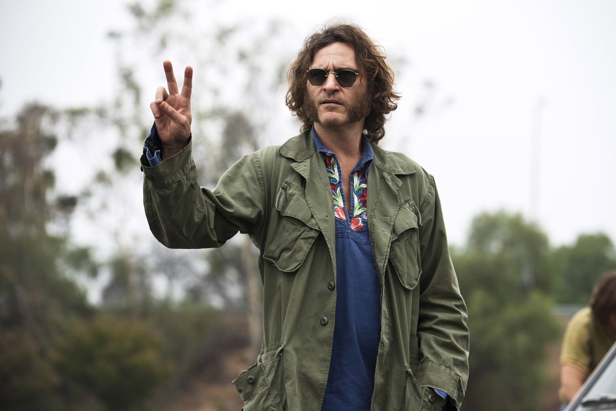 Joaquin Phoenix as Doc Sportello in Inherent Vice wearing a green army jacket and sunglasses and holding up a peace sign