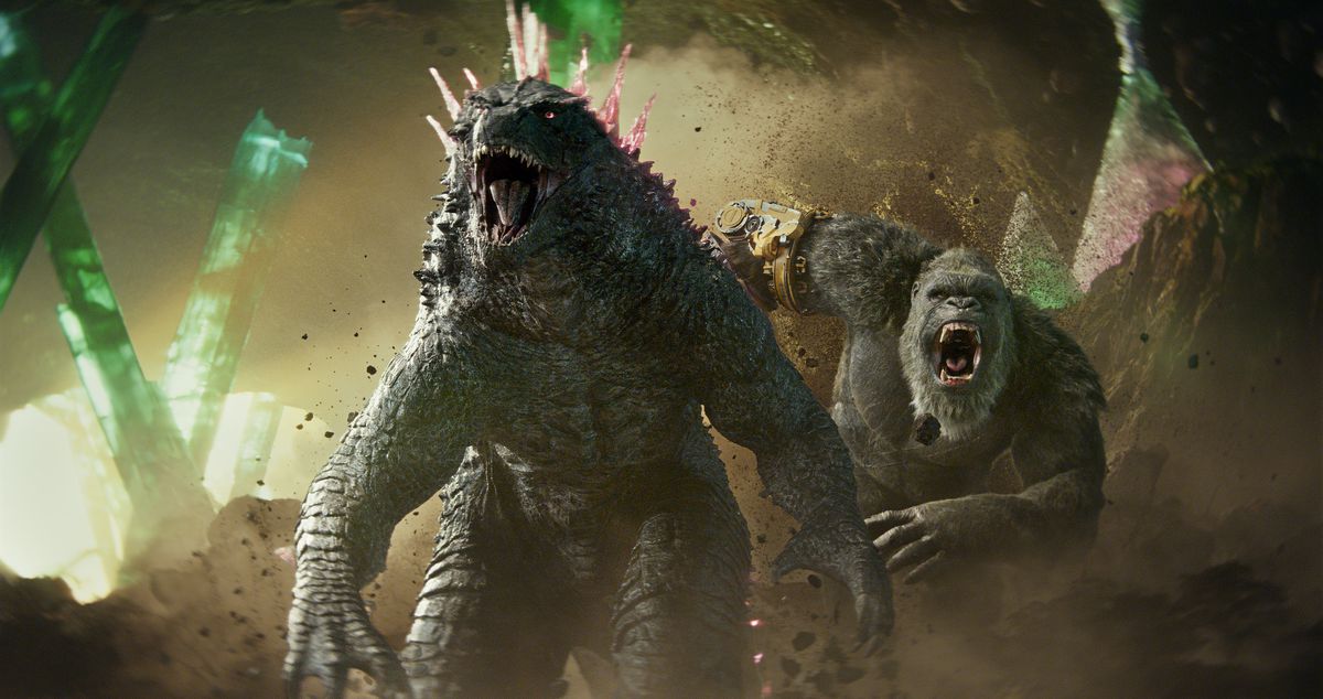 Godzilla, nu med rosa spikar på ryggen, vrålar när King Kong, nu med en robothand, ryter upp bakom honom när de står i en grotta tillsammans i Godzilla x Kong: The New Empire