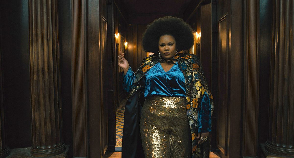 Dede (Nicole Byer), en svart kvinna med en stor, formad afro, iklädd lamébyxor i guld, en elektrisk blå sidenskjorta och en kimonoliknande sidenblommig jacka, poserar dramatiskt i en mörk hall i The American Society of Magical Negroes