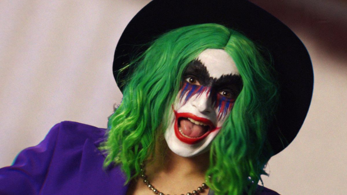 Vera Drew som Joker the Harlequin, en mashup av Joker och Harley Quinn, i grön peruk, clownmålning med ett stort rött läppstiftsleende, en lila kostymjacka och svart hatt i The People's Joker
