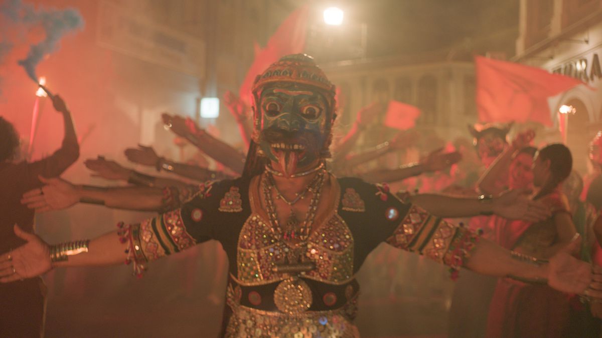 Karaktärer som bär masker och traditionell klädsel på en gatufest håller upp armarna i ett mönster som den hinduiska gudinnan Shiva i Monkey Man