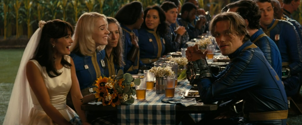 Ella Purnell, iklädd brudklänning, skrattar medan resten av bröllopsfesten pratar vid ett bord i Fallout