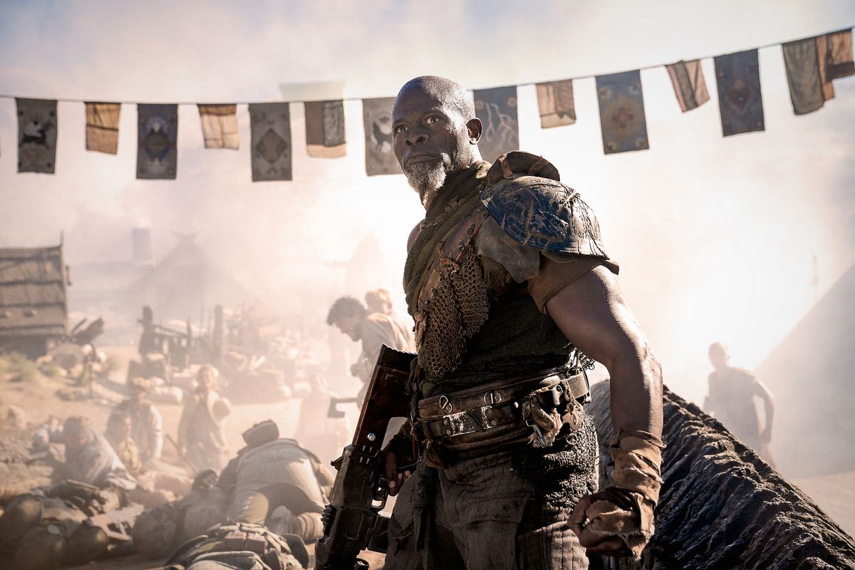 Djimon Hounsou som general Titus i Rebel Moon — Del två: Scargiveren håller en pistol vid sin sida när han står nära en massakrerad by