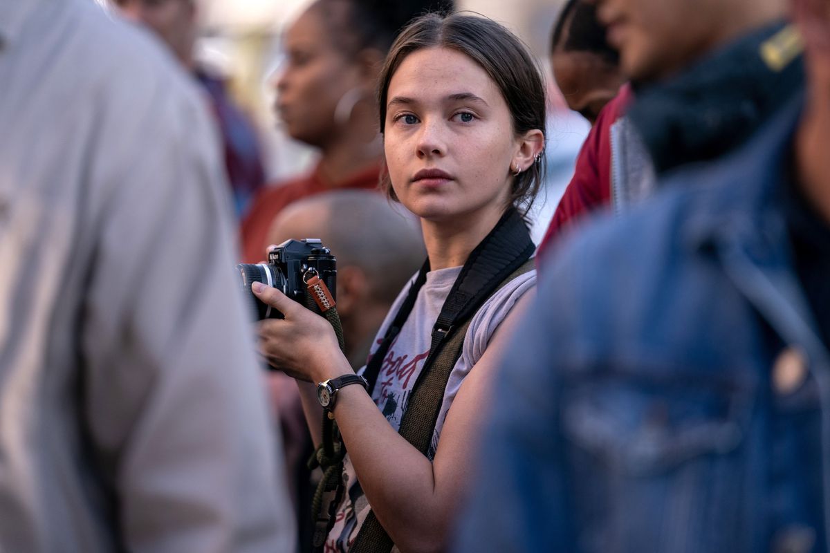 Jessie (Cailee Spaeny), en ung fotojournalist som håller i en kamera, står mitt i en folkmassa i Alex Garlands inbördeskrig