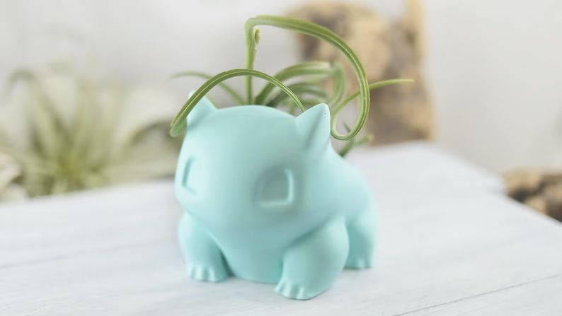 A photo of a teal planter shaped like a Bulbasaur with a succulent planted inside