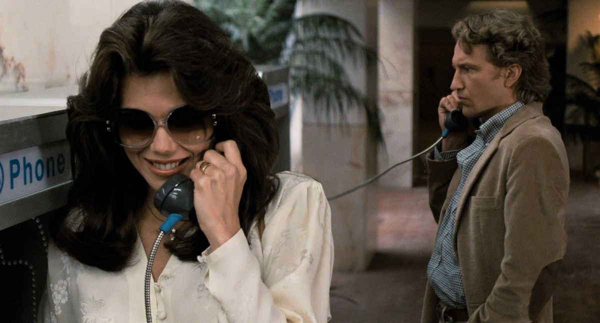 En kvinna som bär solglasögon står vid en telefonkiosk medan en man i brun jacka står i bakgrunden och håller en telefon mot örat.