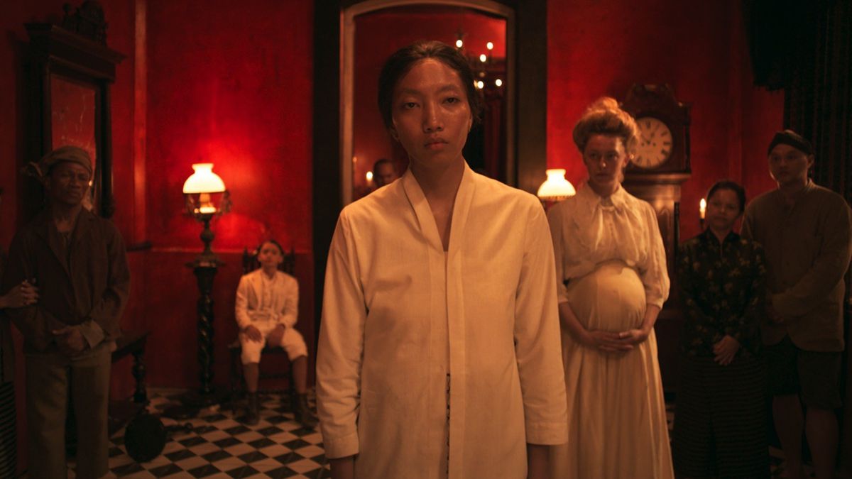 A scene inside a Dutch East Indies plantation house in Sweet Dreams, with a depressed-looking Indonesian woman walking in front of a pregnant white woman in a room with bright red walls as other people look on