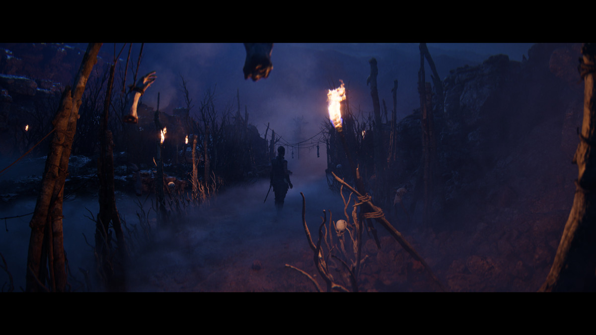 Senua, sedd på avstånd i siluett, utforskar ett mörkt, dimmigt område i Hellblade 2. Avhuggna händer och skallar hänger från grenar.