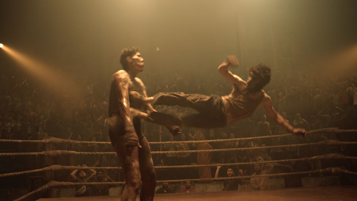 En man i en apmask slår en annan, enorm man med en flygande spark med båda fötterna i en boxningsring i Monkey Man