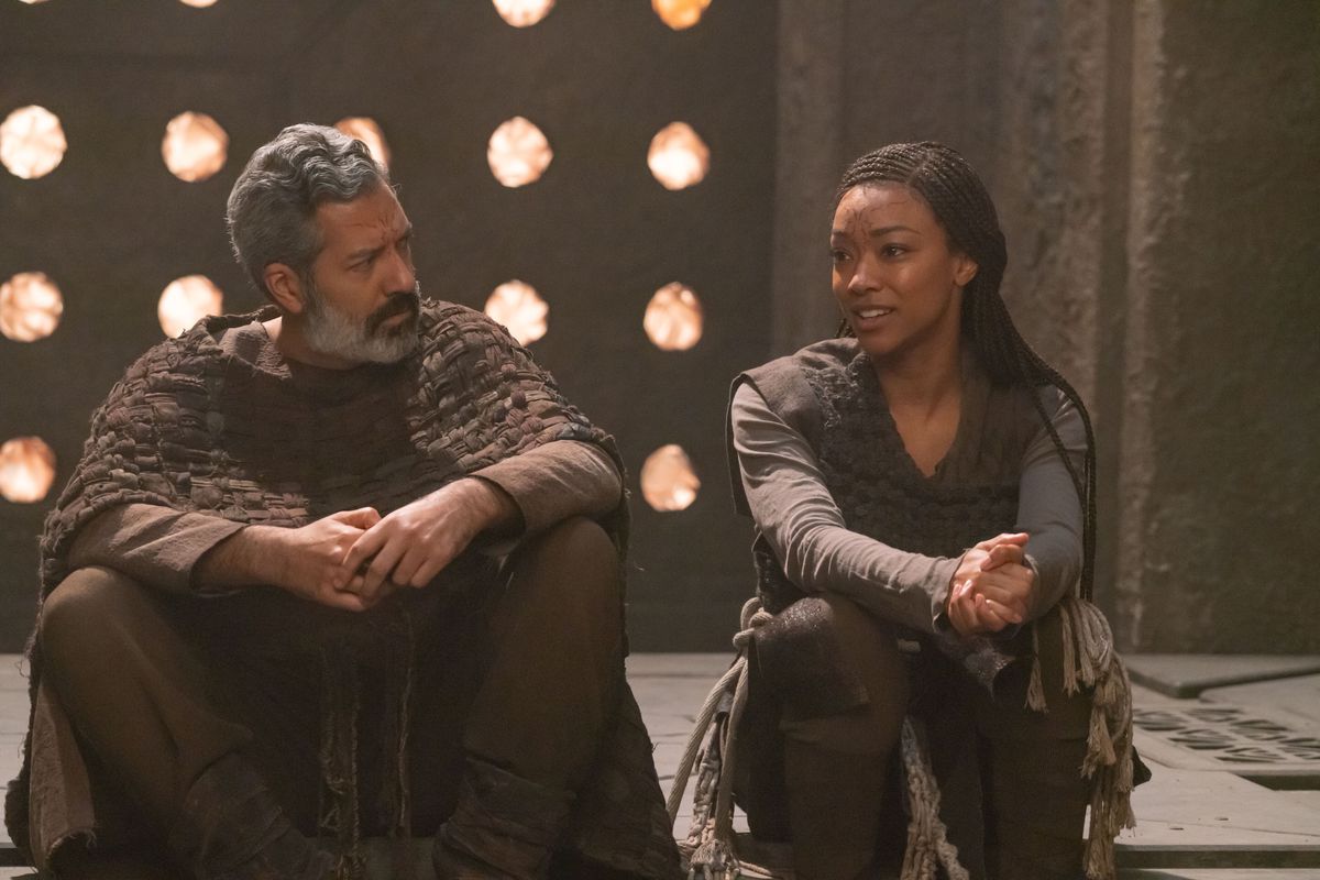 LR Alfredo Narciso som Ohvahz och Sonequa Martin-Green som Michael Burnham i Star Trek: Discovery.  De bär handgjorda utomjordiska plagg och samtalar lugnt medan de sitter på golvet i ett stenrum.