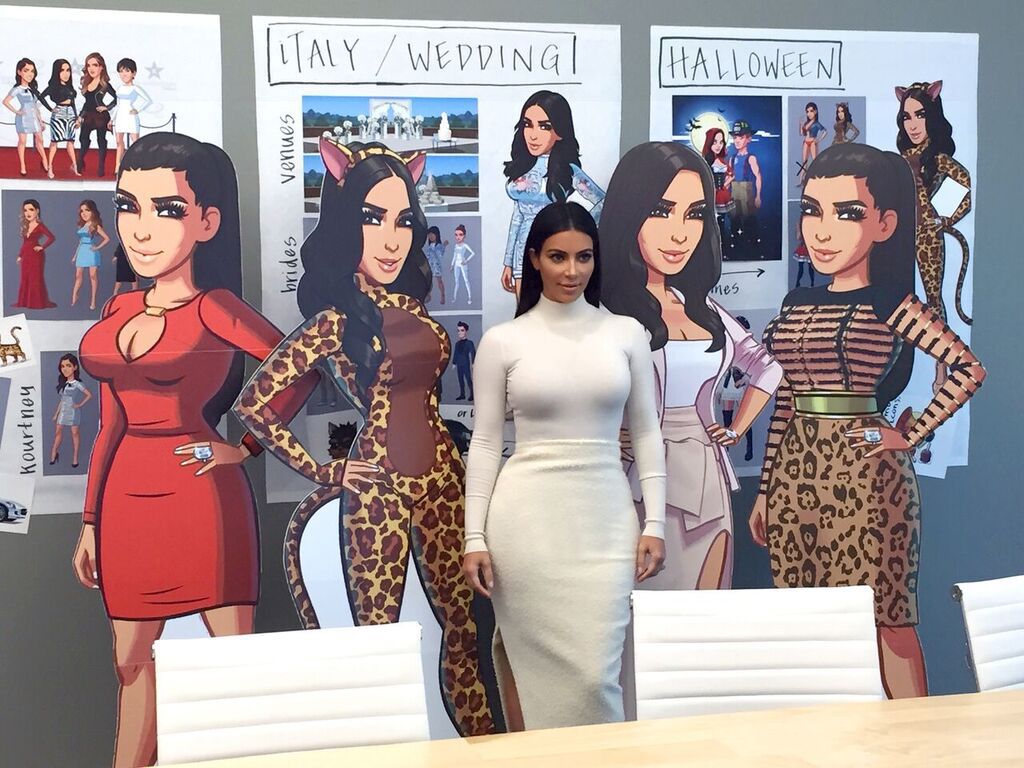 Kim Kardashian wearing a white turtleneck dress standing in front of cardboard cutouts of her character in Kim Kardashian: Hollywood