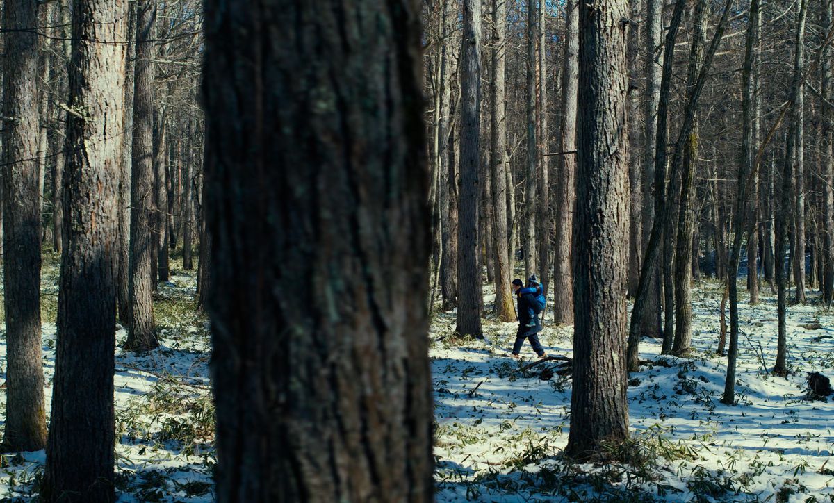 Takumi (Hitoshi Omika) carries his young daughter Hana (Ryo Nishikawa) through a snowy forest on a piggyback ride in an extreme long shot in Ryûsuke Hamaguchi’s Evil Does Not Exist