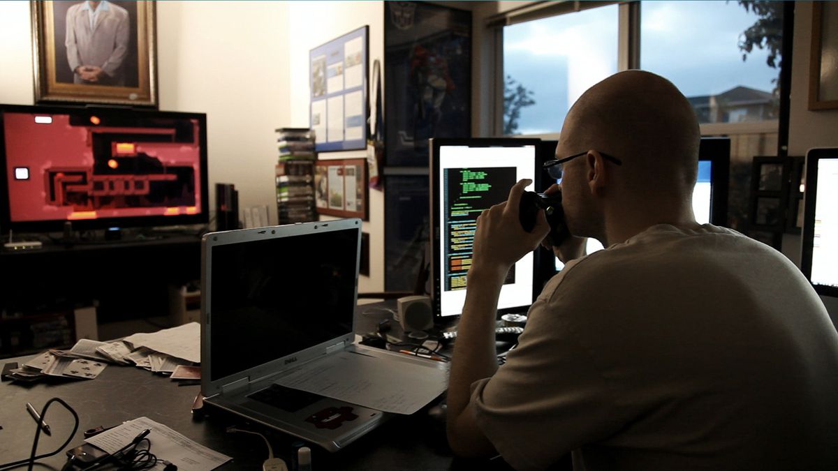 Tommy Refenes sitter vid sitt skrivbord omgiven av datorskärmar och spelar Super Meat Boy på en TV i bakgrunden