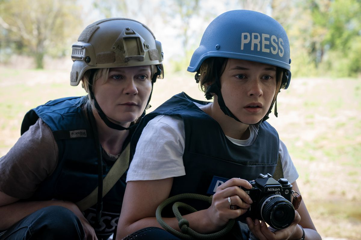 Fotojournalisterna Lee Miller (Kirsten Dunst) och Jessie (Cailee Spaeny) kurar ihop sig, Jessie håller i kameran och bär en klarblå hjälm som säger 