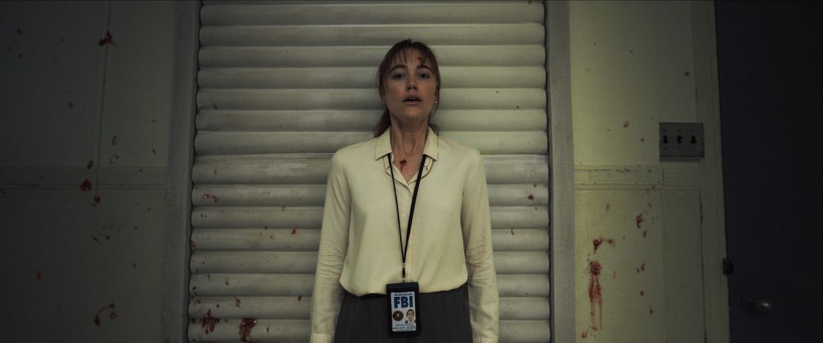 A woman wearing an FBI ID lanyard stands in what seems to be a dim, blood-splattered room in Longlegs.