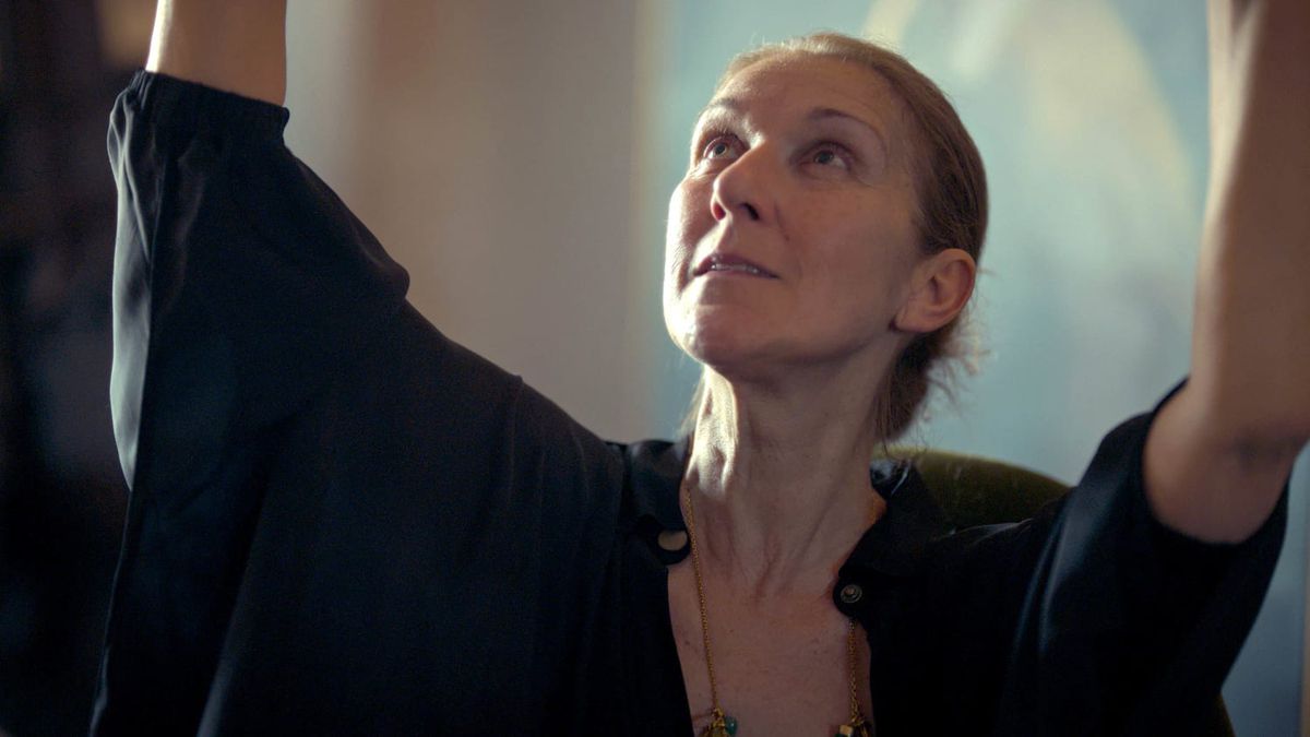 A close-up of a woman (Celine Dion) in a dark shirt raising her arms above her head and looking up in I Am: Celine Dion.