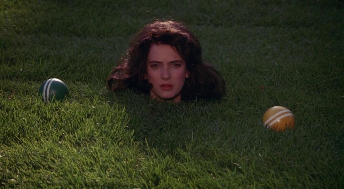 A woman with curly brown hair with her head sticking out of a green lawn with green and yellow polo balls beside her in Heathers.