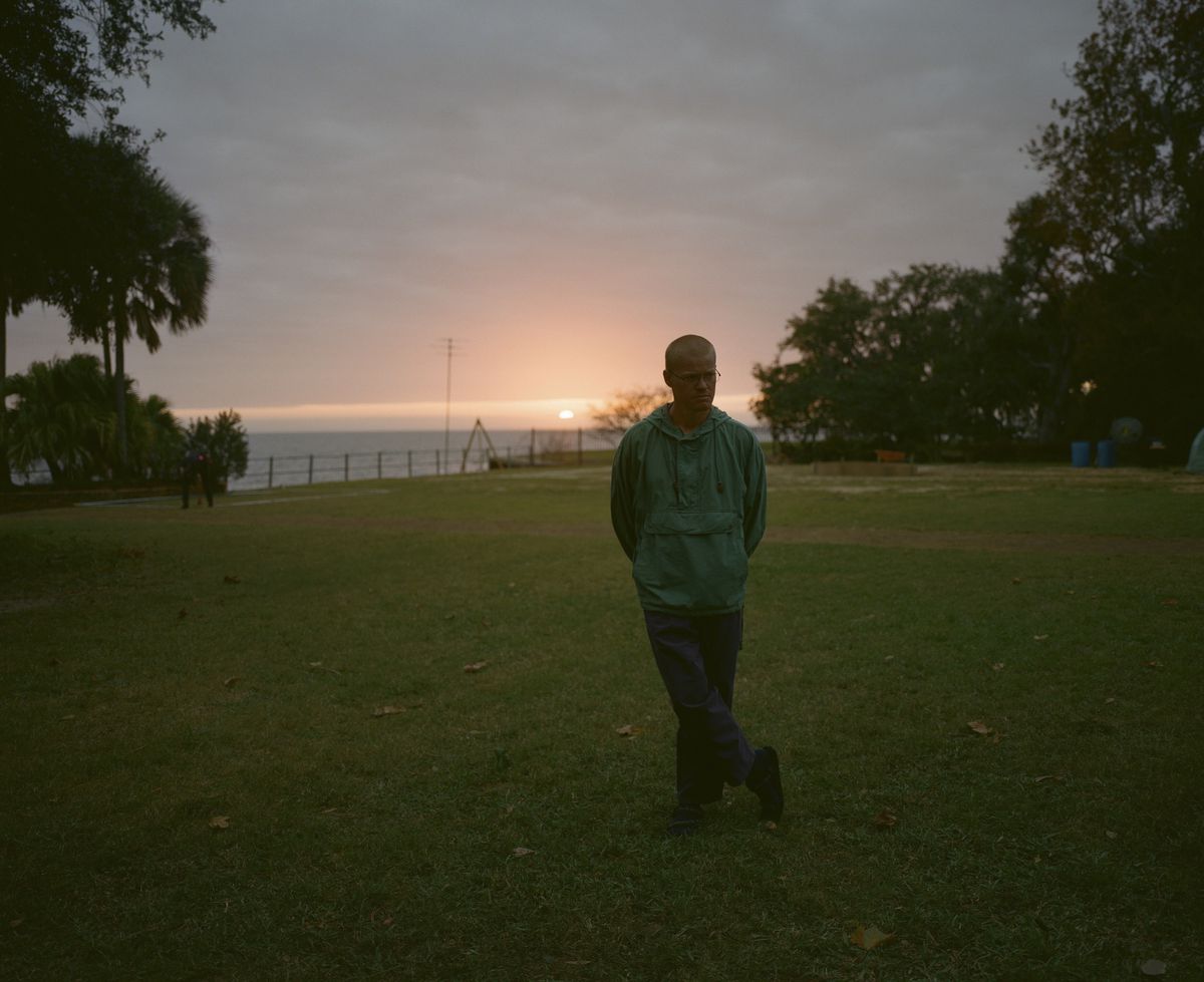 Jesse Plemons, med ett surrsnitt och klädd i en krickfärgad vindjacka, står i en trädgård vid havet i solnedgången i Kinds of Kindness