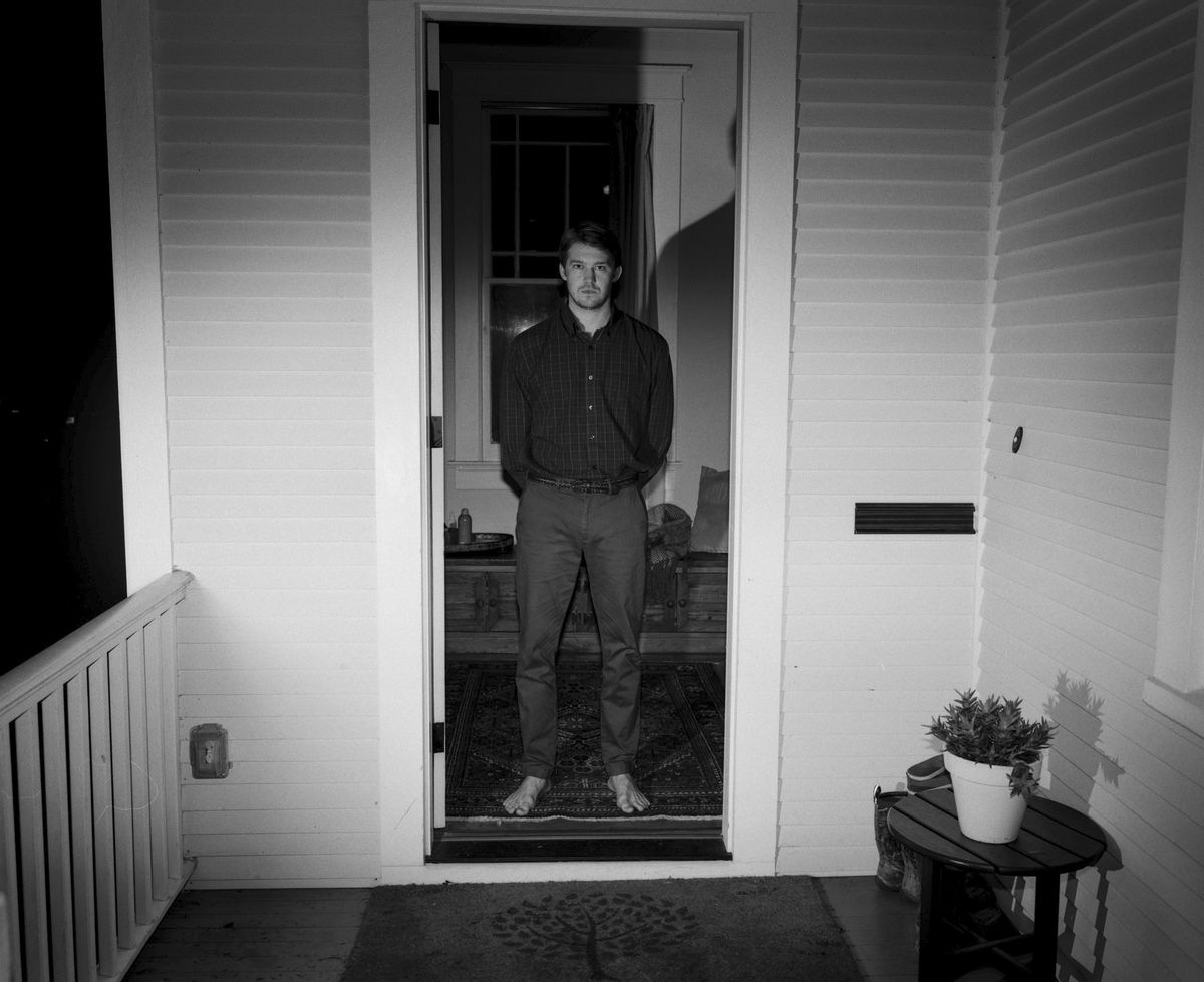 Joe Alwyn, barfota och skjuten i svart och vitt, står i dörröppningen till ett hem i Kinds of Kindness