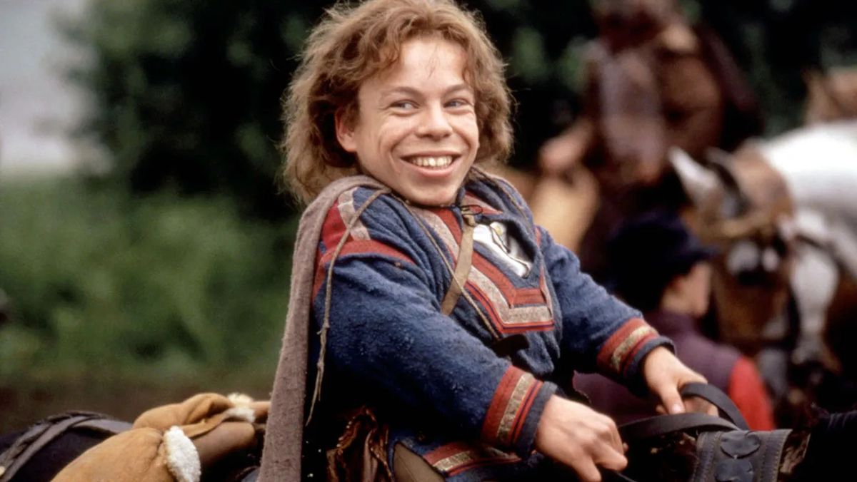 A smiling man (Warwick Davis) seated in the saddle of a horse holding reins in Willows.