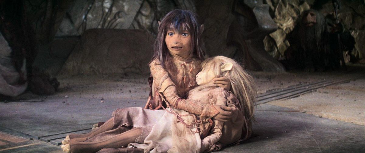 A long-haired male puppet holding the body of a female puppet in a room covered in dirt and rubble in The Dark Crystal.