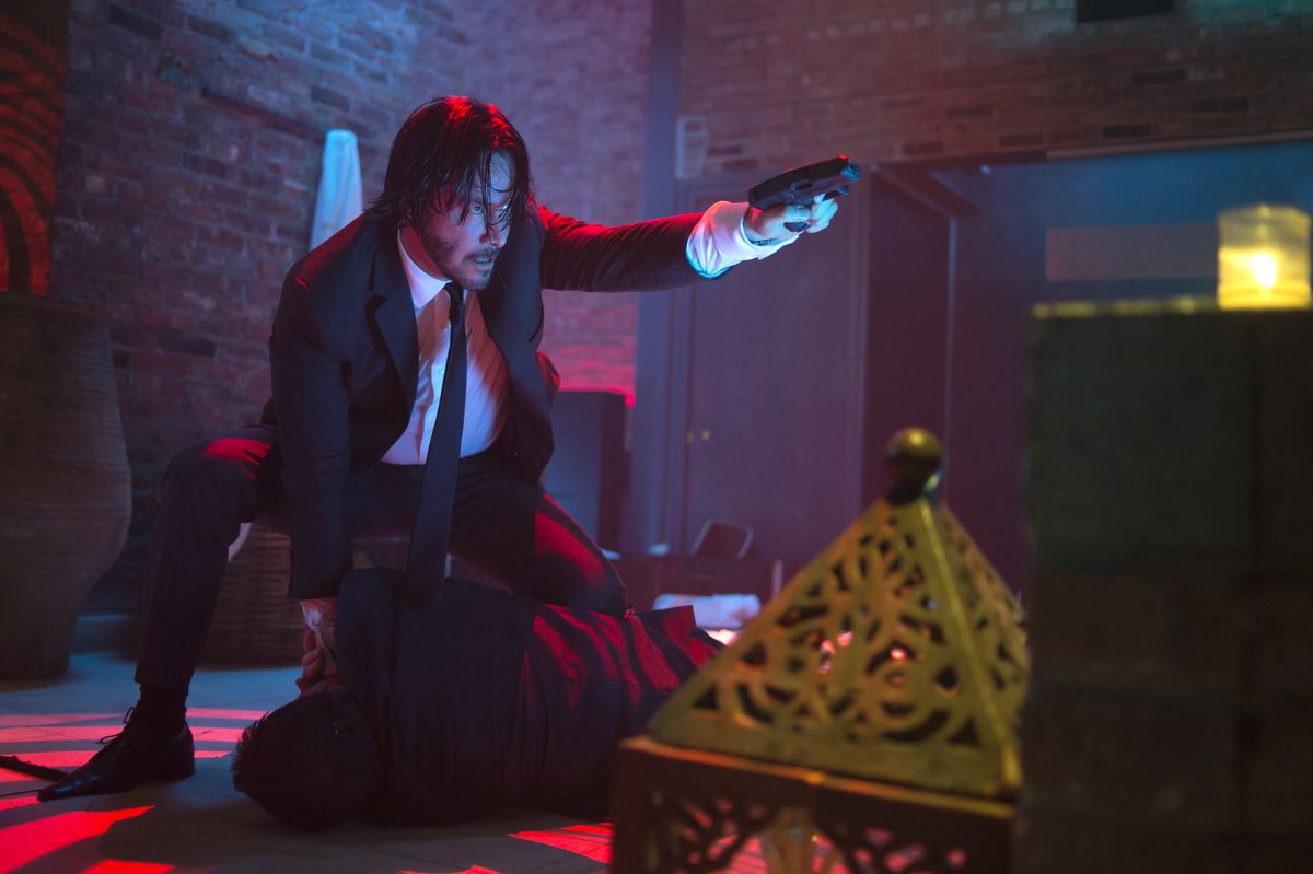 Keanu Reeves, as John Wick in John Wick (2014), aims a gun with his left hand while his right hand is tied up with a body on the floor