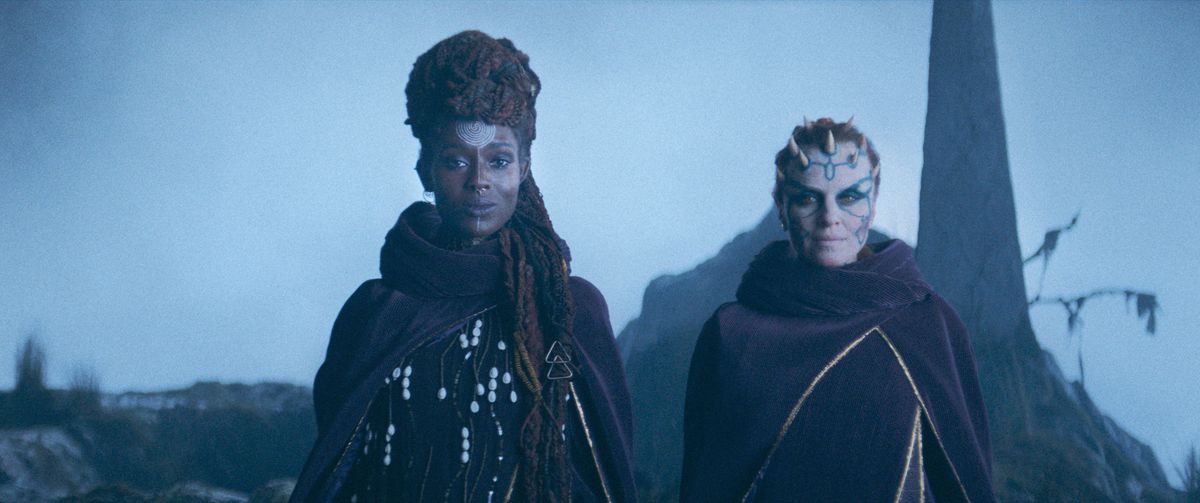 (LR): Mother Aniseya (Jodie Turner-Smith) och Koril (Margarita Levieva) i The Acolyte.  De står bredvid, ler lätt, i utarbetade dräkter på en skarp klippvägg.